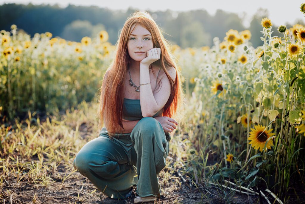 Teen girl with long red hair kneeling in a field of sunflowers at golden hour — a radiant moment from her 10-year senior photo journey in Sarasota.