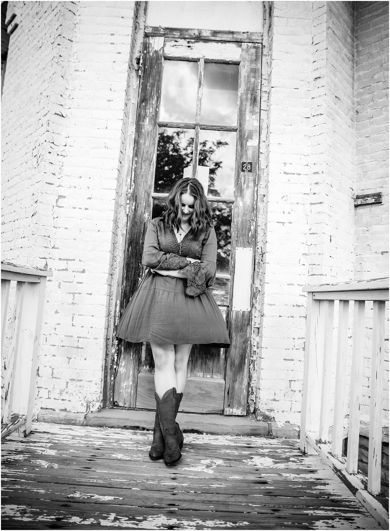 Black and white image of teen girl in a dress and cowboy boots in front of a distressed doorway