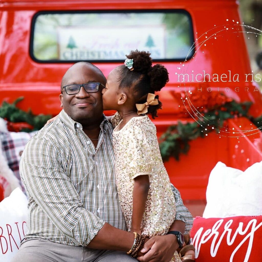 Young girl kissing her father on the cheek while sitting by a vintage red truck decorated for Christmas during Sarasota Holiday Mini Sessions