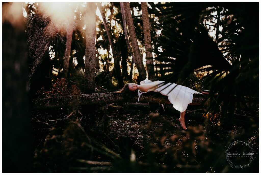 Sarasota senior girl in a white dress lying on a branch barefoot during sunset.