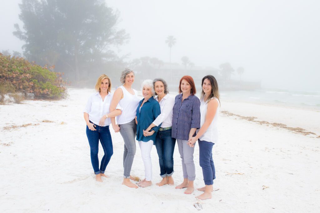 Group of women from different generations laughing and holding hands on a foggy Sarasota beach during a friendship and family photography session.