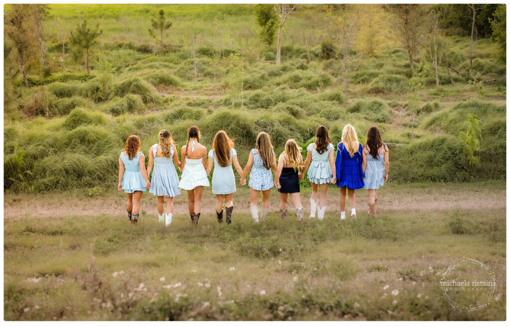 Group of teen girls holding hands and walking away through a grassy field during a golden-hour friendship photography session in Sarasota, Florida, wearing coordinating dresses and boots.