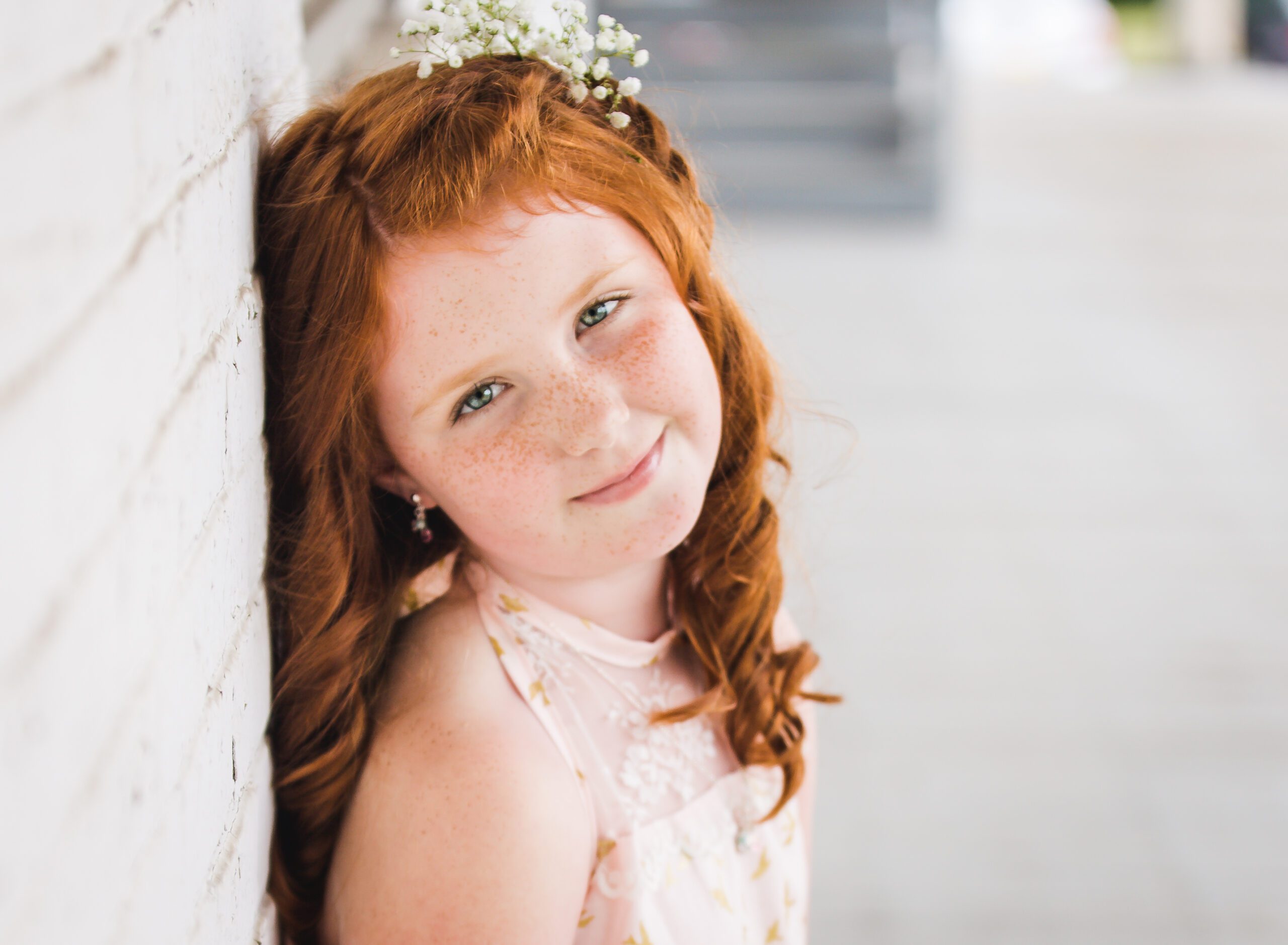 Young girl with red hair and freckles leaning against a white brick wall, smiling softly — an early portrait in a 10-year senior photo journey in Sarasota.