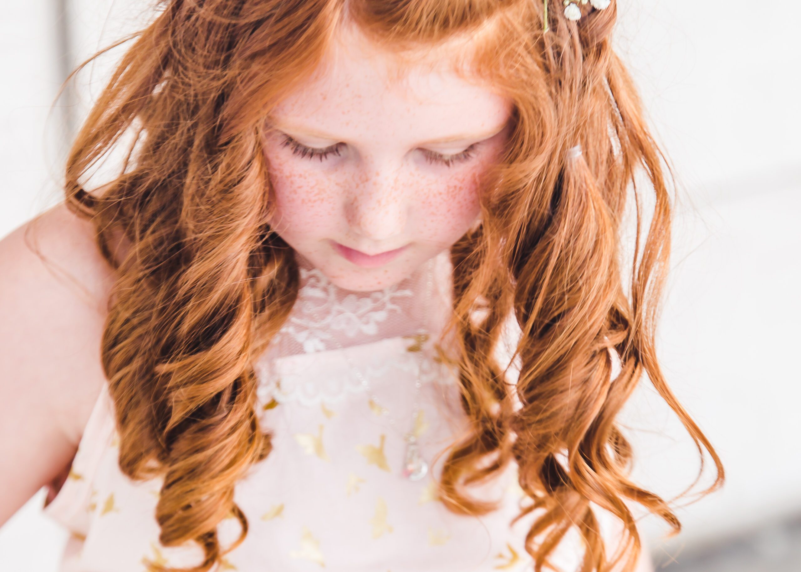 Young redheaded girl with curled hair looking down thoughtfully in a soft pink and gold dress — an early moment from a 10-year senior photo journey in Sarasota.
