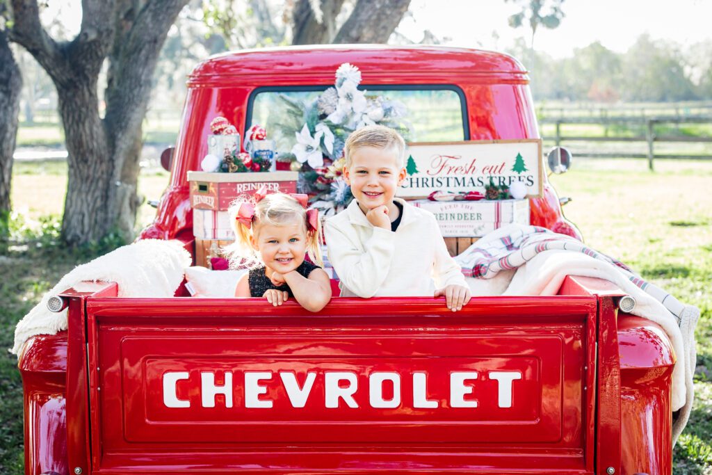 Brother and sister smiling while sitting in the back of a decorated vintage red Chevrolet truck during Sarasota Holiday Mini Sessions