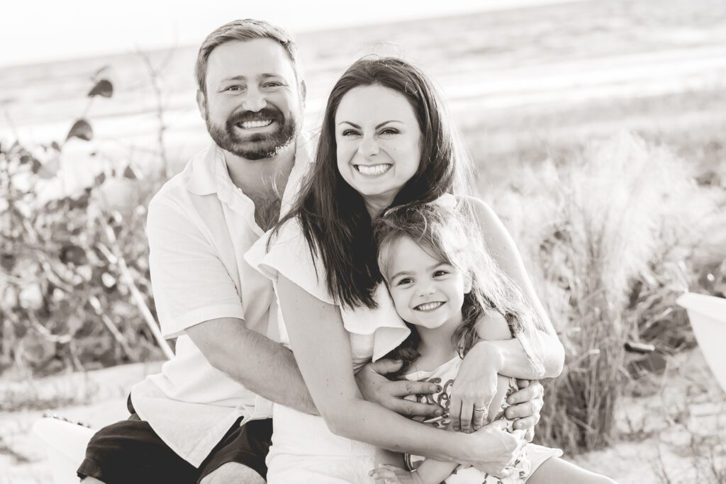Smiling family of three sitting together on the beach during a Sarasota photo session, black and white portrait.