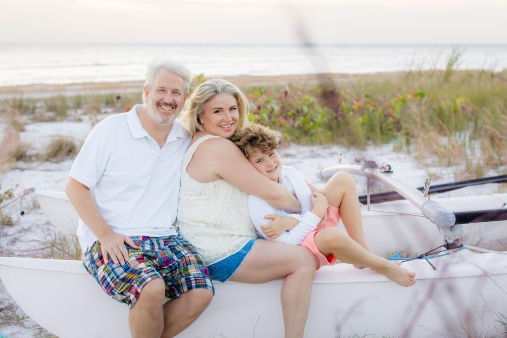 Family sitting together on a sailboat at the beach during a Sarasota photo session — smiling parents with their young son at sunset.