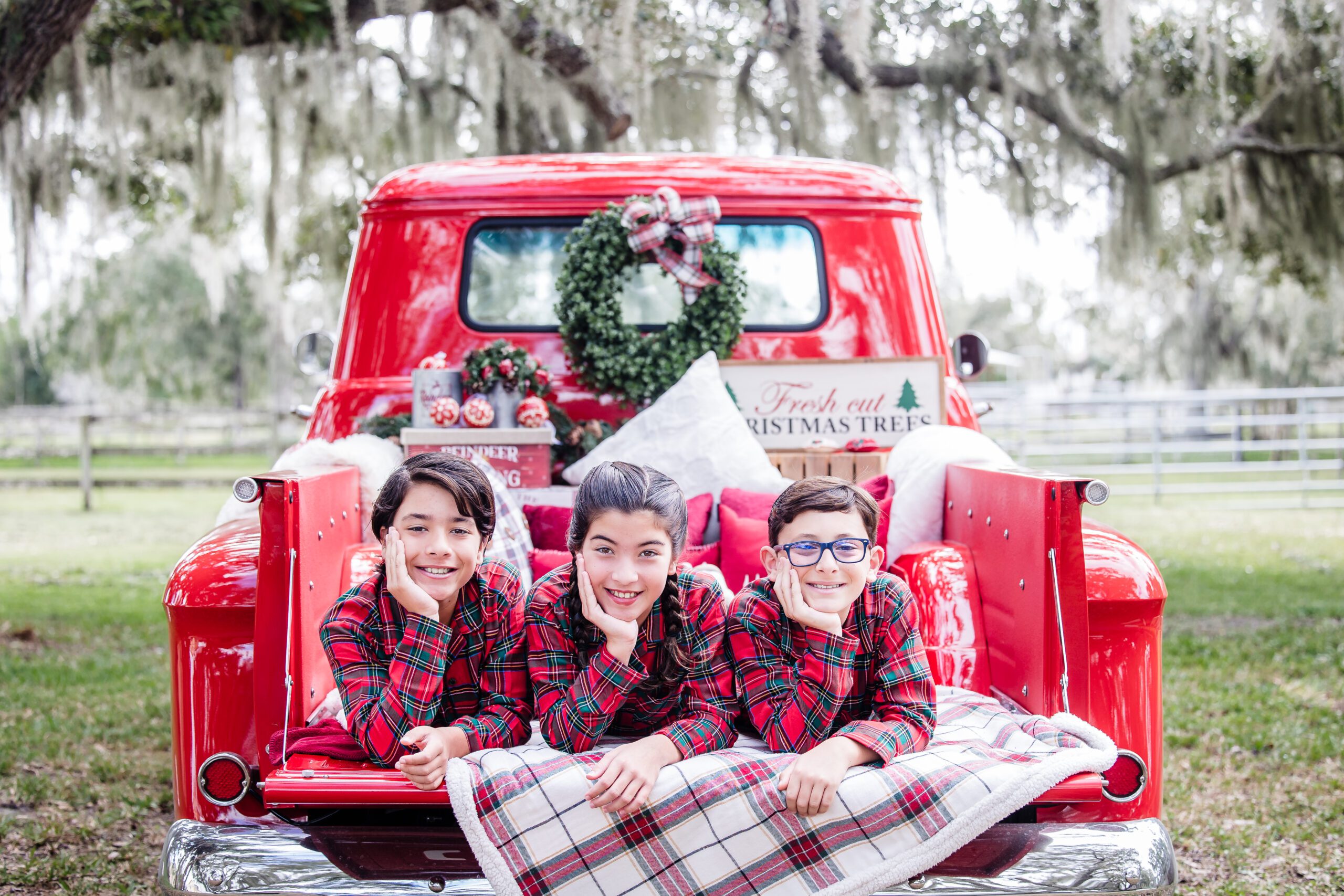 Three kids in matching red plaid pajamas posing on the back of a vintage red truck decorated for Christmas during a Sarasota holiday mini session
