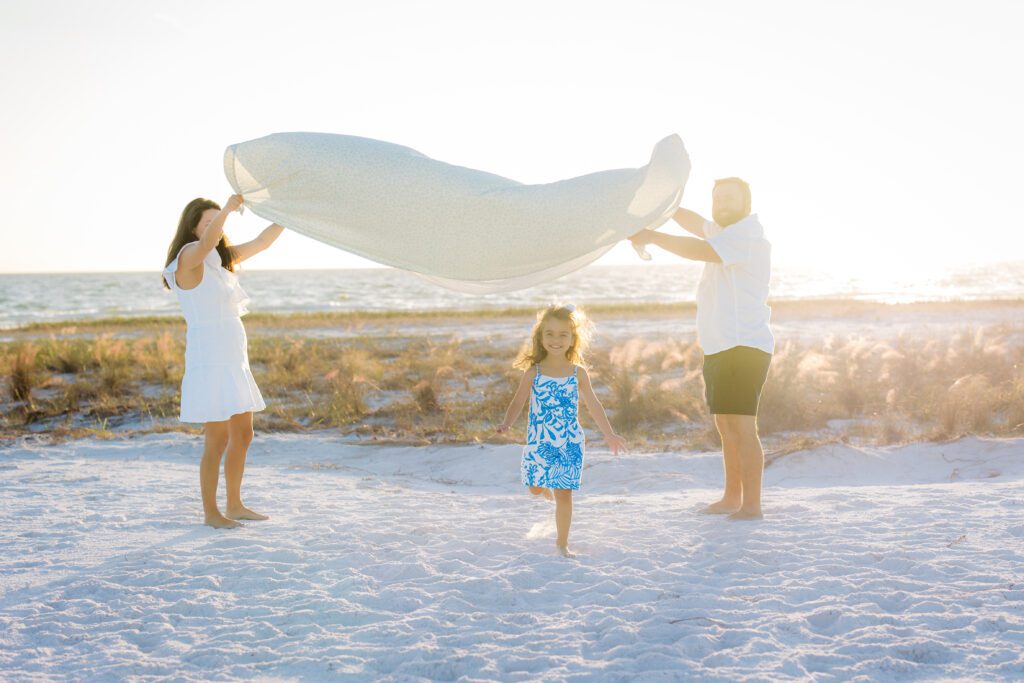 Little girl giggling as she runs under a white bedsheet held up by parents at Siesta Key Beach during family photo mini session.
