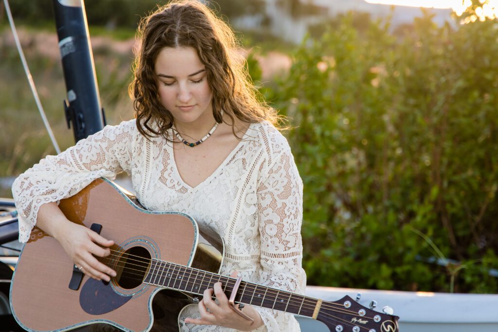 High school senior girl playing guitar during sunrise photo session in Sarasota, Florida, wearing a lace dress with golden light behind her