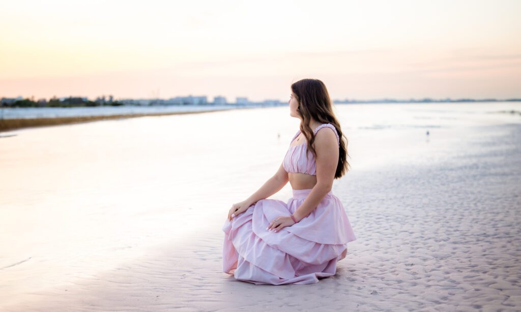 High school senior girl in a pink dress kneeling on the beach at sunrise in Sarasota, Florida, looking toward the water with soft pastel skies behind her.
