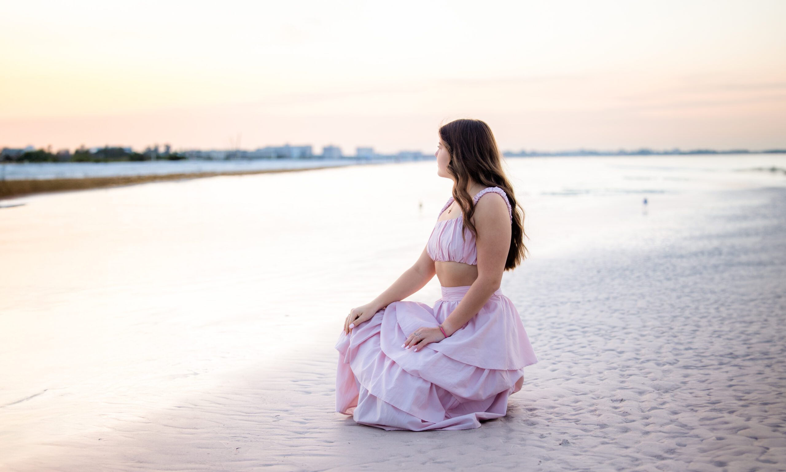 High school senior girl in a pink dress kneeling on the beach at sunrise in Sarasota, Florida, looking toward the water with soft pastel skies behind her.