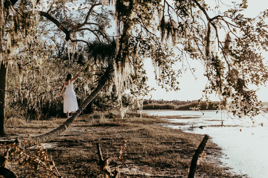 High school senior in white dress balancing on fallen tree at Myakka River State Park with Spanish moss and river in background during Sarasota senior portrait session
