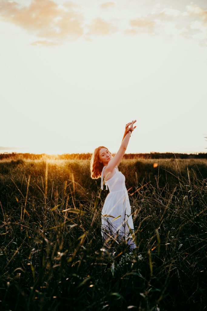 High school senior in white dress with arm raised standing in golden hour prairie field at Myakka River State Park during Sarasota senior portrait session
