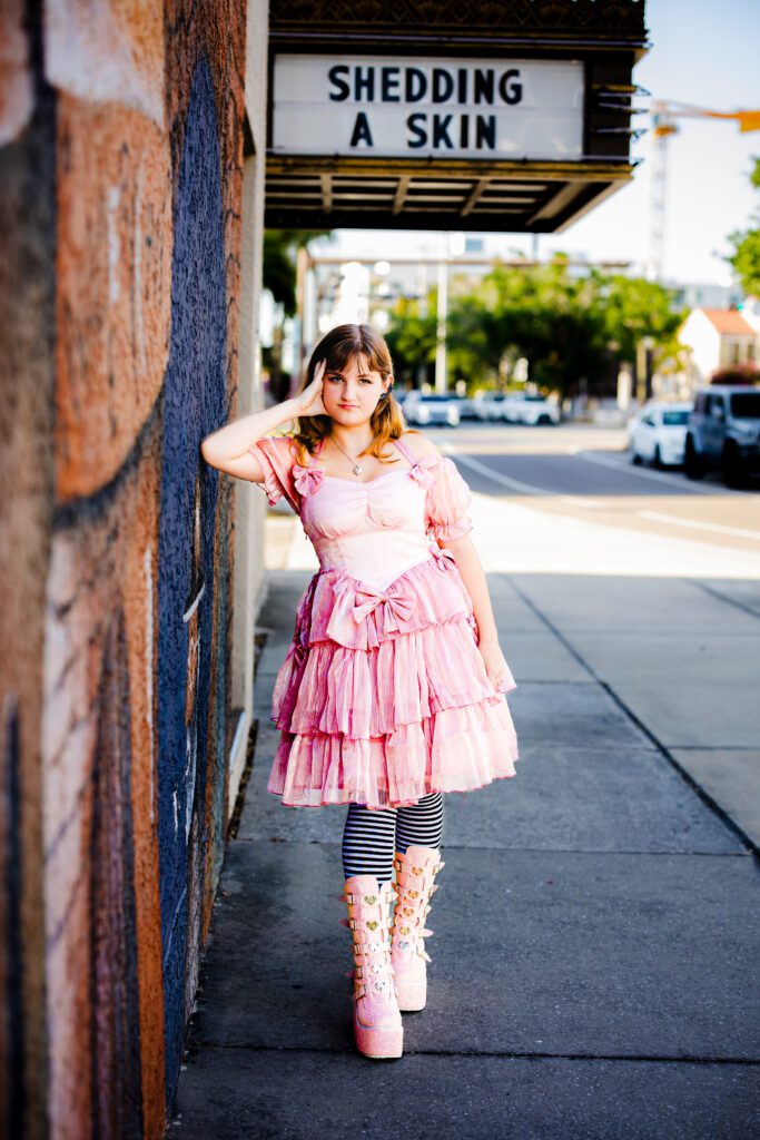 High school senior in pink ruffle dress and platform boots standing in front of Shedding a Skin marquee in downtown Sarasota during senior portrait session
