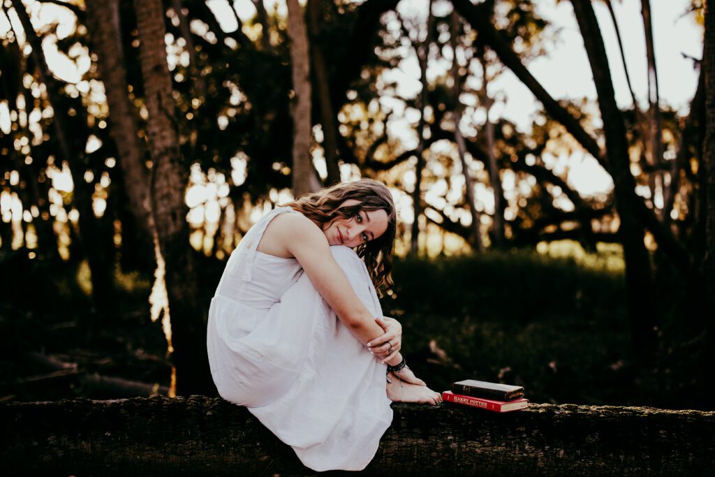 High school senior in white dress sitting on mossy log with Harry Potter books at Myakka River State Park during Sarasota senior portrait session