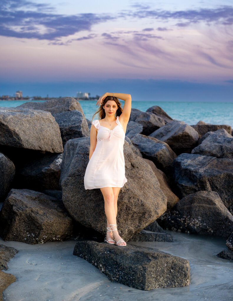 Senior portrait on the beach at sunset in Sarasota Florida, girl in pink dress standing on rocks with lavender sky over the Gulf of Mexico, Michaela Ristaino Photography