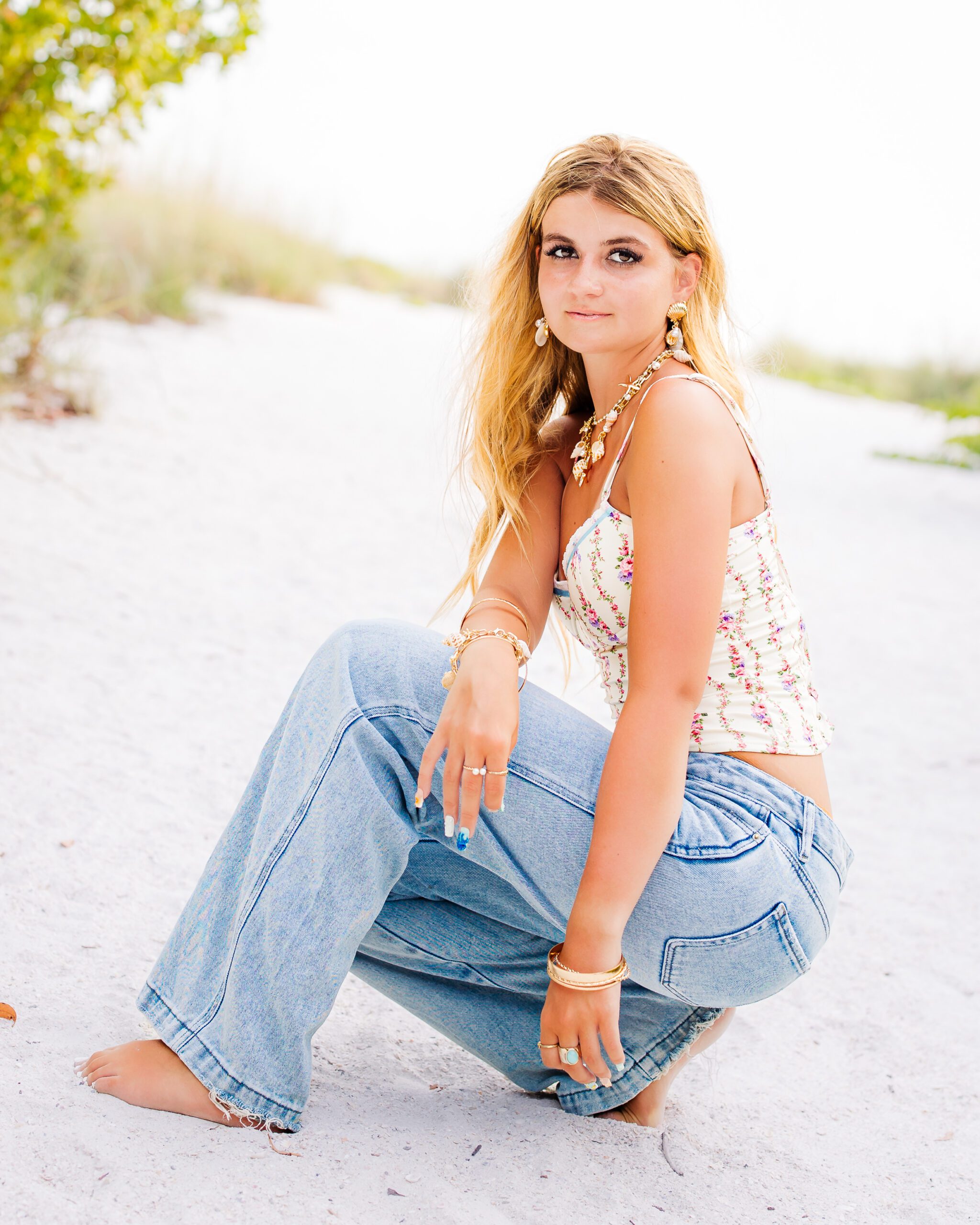 What to wear for senior photos: Sarasota senior girl in floral top and jeans crouching on the beach, layered gold jewelry and rings
