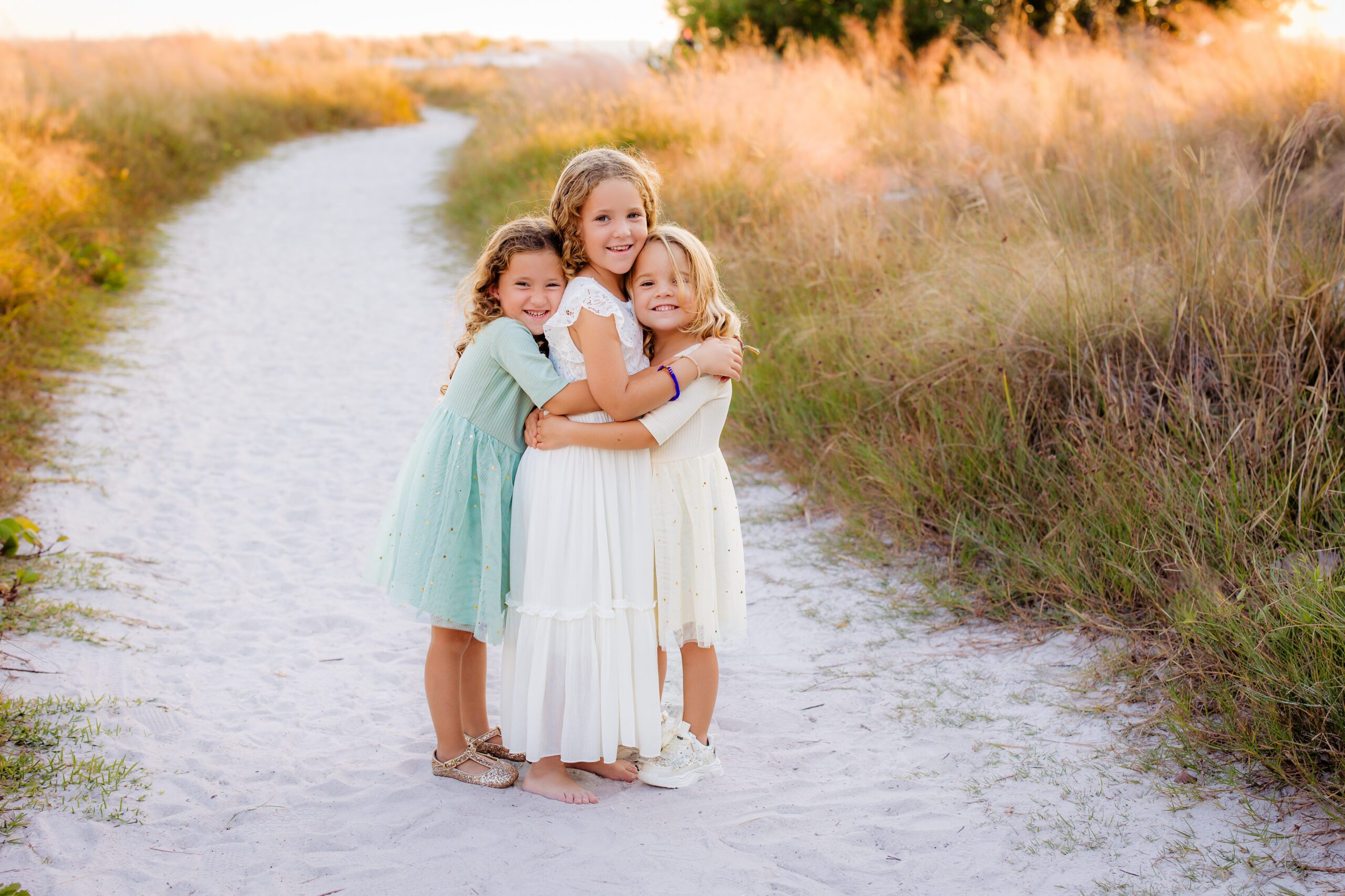 Three sisters hugging and laughing on a sandy path during a Sarasota Holiday Mini Session